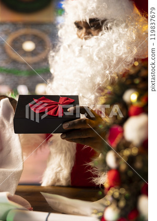 Father Christmas prepares a beautifully wrapped gift box in his workshop, using bows and wrapping paper. Santa spreading the festive holiday spirit during winter, x mas magic and fantasy. Close up. 119837679