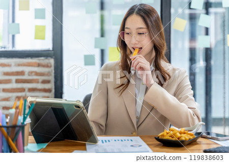 Young businesswoman eating unhealthy snack, french fried, while working on her project at her desk 119838603