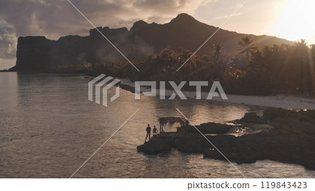 Two tourists standing on rocks shore, enjoy breathtaking bright sunset over mountain silhouette in Maupiti, palm trees lining white sand beach. People enjoy remote wild nature summer vacation travel Two tourists standing on rocks shore, enjoy breathtaking bright sunset over mountain silhouette in Maupiti, palm trees lining white sand beach. People enjoy remote wild nature summer vacation travel 119843423
