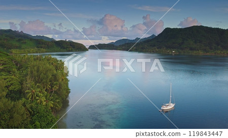 Aerial drone footage captures a serene sailboat anchored in the crystal-clear waters of Opunohu bay, Moorea, as the sun sets, casting a warm glow over the lush tropical landscape and tranquil bay 119843447
