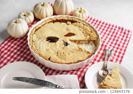 Halloween food. Homemade pumpkin pie or tart with a scary face for Halloween on a red napkin on gray table with cut piece on the plate. Copy space. Halloween food concept. 119843576