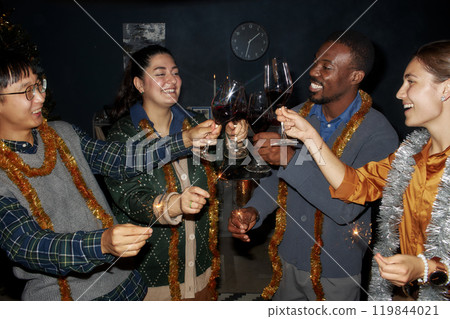 Medium shot of diverse group of cheerful friends wearing vintage outfits and tinsels clinking glasses with wine while celebrating Christmas together in living room at home, camera flash Medium shot of diverse group of cheerful friends wearing vintage outfits and tinsels clinking glasses with wine while celebrating Christmas together in living room at home, camera flash 119844021