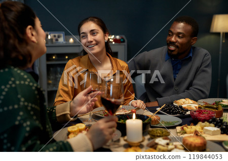 Medium shot of cheerful woman and Black man chatting with female friend sitting at festive dining table celebrating Christmas with friends at home party 119844053