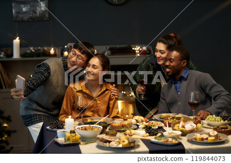 Candid shot of four cheerful friends taking selfie or video calling relatives while sitting at festive dining table celebrating Christmas at home party Candid shot of four cheerful friends taking selfie or video calling relatives while sitting at festive dining table celebrating Christmas at home party 119844073