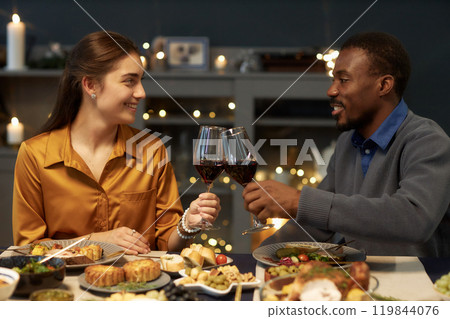 Side view of smiling Black man clinking wine glasses with cheerful woman on romantic date or Christmas celebration while sitting together at dining table in living room, copy space 119844076