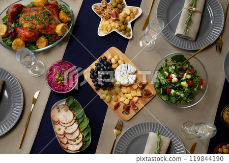 Top view of cheese and fruit platter served on wooden board as appetizer at festive dining table, with simple salads and roasted chicken as main course for Christmas dinner 119844190