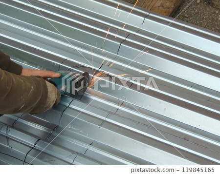 A man in dirty work clothes and gloves cuts a metal sheet with a grinder along the black markings on a corrugated surface, causing sparks to fly. 119845165