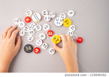 Child hand holding white bottle cap with math problems among scattered numbered caps on gray background, flat lay educational tool for math learning, counting games, interactive math activity kids 119846302
