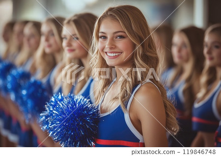 Cheerleader smiling while holding pom poms with teammates in background 119846748