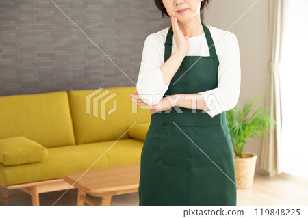 A senior woman housewife/housekeeper wearing a green apron worried in the living room at home A senior woman housewife/housekeeper wearing a green apron worried in the living room at home 119848225