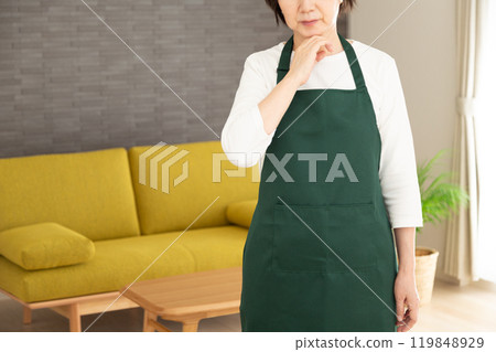 A senior woman housewife/housekeeper wearing a green apron worried in the living room at home A senior woman housewife/housekeeper wearing a green apron worried in the living room at home 119848929