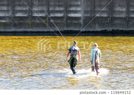Two fishermen wading in shallow river waters near a concrete dam, ready for sturgeon fishing, serene scene blending nature and industrial backdrop 119849152