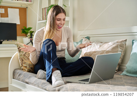 Young happy woman sitting with laptop on couch at home having video call 119854207