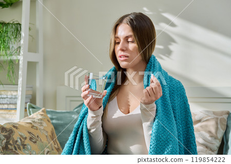 Young woman taking medicine, holding glass of water, sitting on couch at home Young woman taking medicine, holding glass of water, sitting on couch at home 119854222