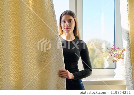 Young beautiful woman looking at camera, with curtain in hand, window background Young beautiful woman looking at camera, with curtain in hand, window background 119854231