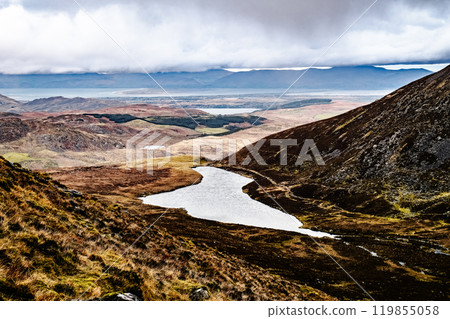 The landscape features a peaceful lake nestled between hills, reflecting the overcast clouds above, with expansive fields leading to distant mountains under a dramatic sky. The landscape features a peaceful lake nestled between hills, reflecting the overcast clouds above, with expansive fields leading to distant mountains under a dramatic sky. 119855058