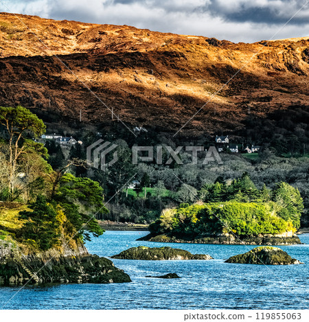 A tranquil bay reveals lush greenery on small islands and dramatic hillsides illuminated by the setting sun, creating a serene atmosphere in Ireland. 119855063