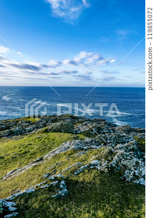 The coastline features dramatic rocky formations and lush greenery, with the ocean stretching far into the horizon under a bright blue sky during early morning. The coastline features dramatic rocky formations and lush greenery, with the ocean stretching far into the horizon under a bright blue sky during early morning. 119855072