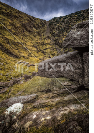 A rugged path winds through a lush, mossy landscape, flanked by rocky outcrops under a moody, overcast sky. Nature reveals its beauty in every detail. 119855077