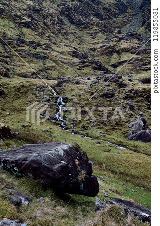 A gentle stream meanders through a rocky valley, bordered by vibrant green grass and scattered boulders. 119855081