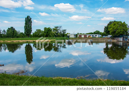 The river flows through the city. On the calm surface of the river banks are reflected. Slovak river Nitra on a bright sunny day. 119855184