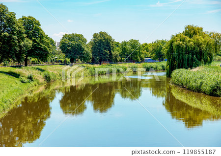 The river flows through the city. On the calm surface of the river banks are reflected. Slovak river Nitra on a bright sunny day. The river flows through the city. On the calm surface of the river banks are reflected. Slovak river Nitra on a bright sunny day. 119855187