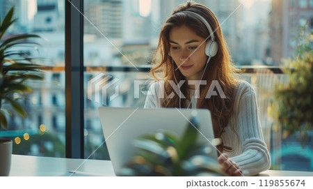 A businesswoman with headphones working on a laptop in a stylish, modern office space with large windows and city views. A businesswoman with headphones working on a laptop in a stylish, modern office space with large windows and city views. 119855674