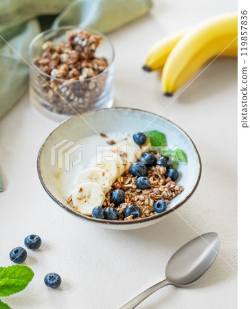 Granola with yogurt, banana and blueberries in a bowl on a light background with spoon and napkin. 119857836