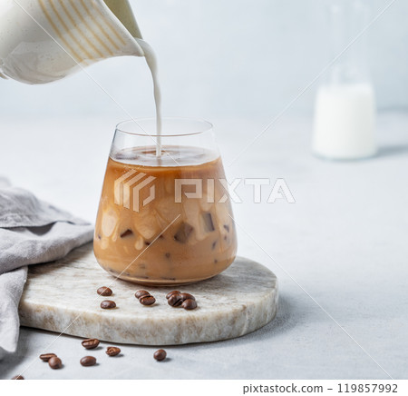 Milk pouring from a milk jug into a glass of iced coffee on a marble board on a light background Milk pouring from a milk jug into a glass of iced coffee on a marble board on a light background 119857992