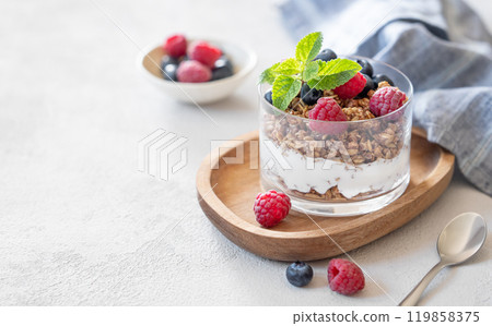Natural yogurt with granola, raspberries and blueberries in a glass on a wooden board 119858375