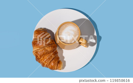 Flat lay of coffee cup and fresh croissant on a white plate on a blue background. Flat lay of coffee cup and fresh croissant on a white plate on a blue background. 119858616
