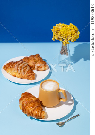 Coffee cup and fresh croissants on blue background with yellow flowers and shadow. Coffee cup and fresh croissants on blue background with yellow flowers and shadow. 119858618