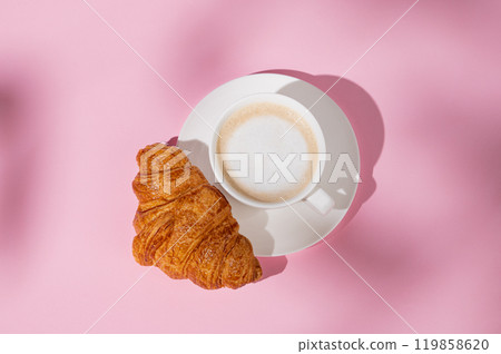 Flat lay of coffee cup and fresh croissant on a white plate on a pink background with shadow. 119858620