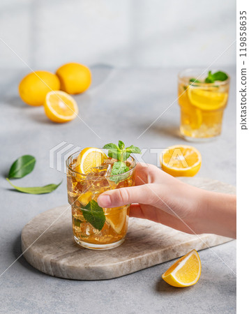 A hand takes a glass of iced tea with lemon and mint against a gray background with citrus fruits A hand takes a glass of iced tea with lemon and mint against a gray background with citrus fruits 119858635