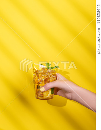 A hand holds a glass of iced tea with lemon and mint on a yellow background with shadow. 119858643
