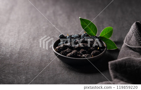 Fresh black mulberry with leaf in black wooden bowl on dark background with napkin. 119859220