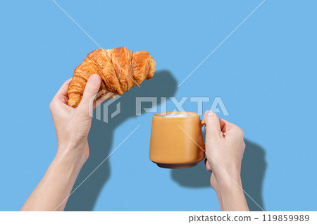 Women's hands hold fresh croissant and cup of coffee  on a blue background.  119859989