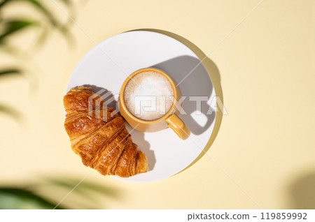 Flat lay of coffee cup and fresh croissant on a white plate on a yellow background Flat lay of coffee cup and fresh croissant on a white plate on a yellow background 119859992