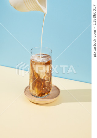 Iced coffee in a tall glass with a milk jug pouring milk on a blue and yellow background with shadow 119860017