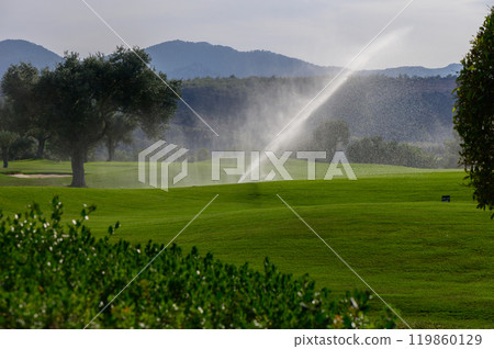 Morning mist rises over a lush golf course as sprinklers create a refreshing spectacle against the backdrop of distant hills 119860129