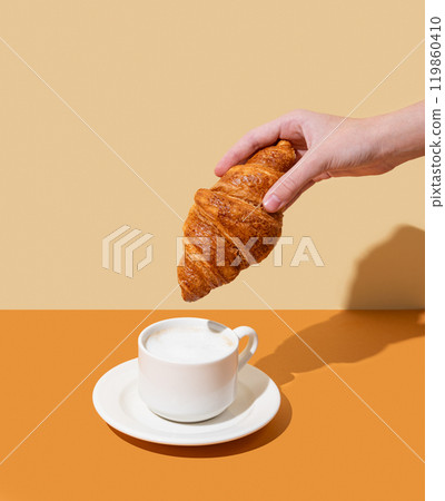 Hand holding fresh croissant over cup with cappuccino coffee on yellow background. Hand holding fresh croissant over cup with cappuccino coffee on yellow background. 119860410