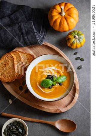 Pumpkin soup with cream, basil and seeds on a wooden board with fried bread on a dark background 119860525