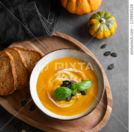 Pumpkin soup with cream, basil and seeds on a wooden board with fried bread on a dark background 119860526