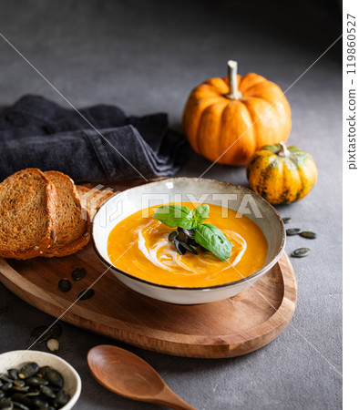 Pumpkin soup with cream, basil and seeds on a wooden board with fried bread on a dark background 119860527
