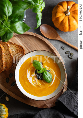 Pumpkin soup with cream, basil and seeds on a wooden board with fried bread on a dark background 119860528
