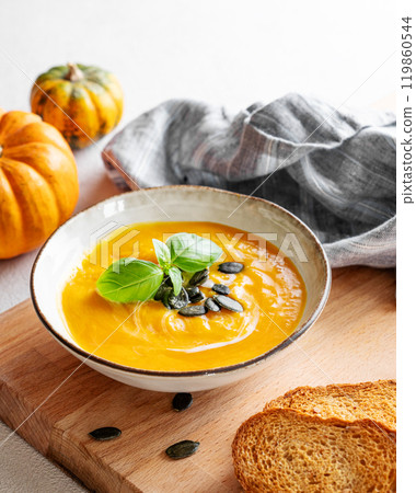 Homemade pumpkin soup with cream, basil and seeds on a wooden board with bread and napkin Homemade pumpkin soup with cream, basil and seeds on a wooden board with bread and napkin 119860544