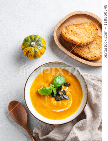 Pumpkin soup with cream, basil and seeds on a light background with bread, spoon and napkin. Pumpkin soup with cream, basil and seeds on a light background with bread, spoon and napkin. 119860549