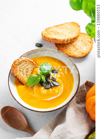 Pumpkin soup with cream, basil and seeds on a light background with bread, fresh pumpkins and spoon. Pumpkin soup with cream, basil and seeds on a light background with bread, fresh pumpkins and spoon. 119860553