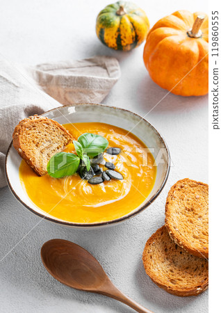 Pumpkin soup with cream, basil and seeds on a light background with bread, spoon and fresh pumpkins. Pumpkin soup with cream, basil and seeds on a light background with bread, spoon and fresh pumpkins. 119860555