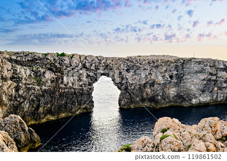 Spectacular sunset at Pont den Gil (Cala Corbetar) in Ciutadella, Menorca, Balearic Islands. Peculiar natural formation in the form of an arch Spectacular sunset at Pont den Gil (Cala Corbetar) in Ciutadella, Menorca, Balearic Islands. Peculiar natural formation in the form of an arch 119861502